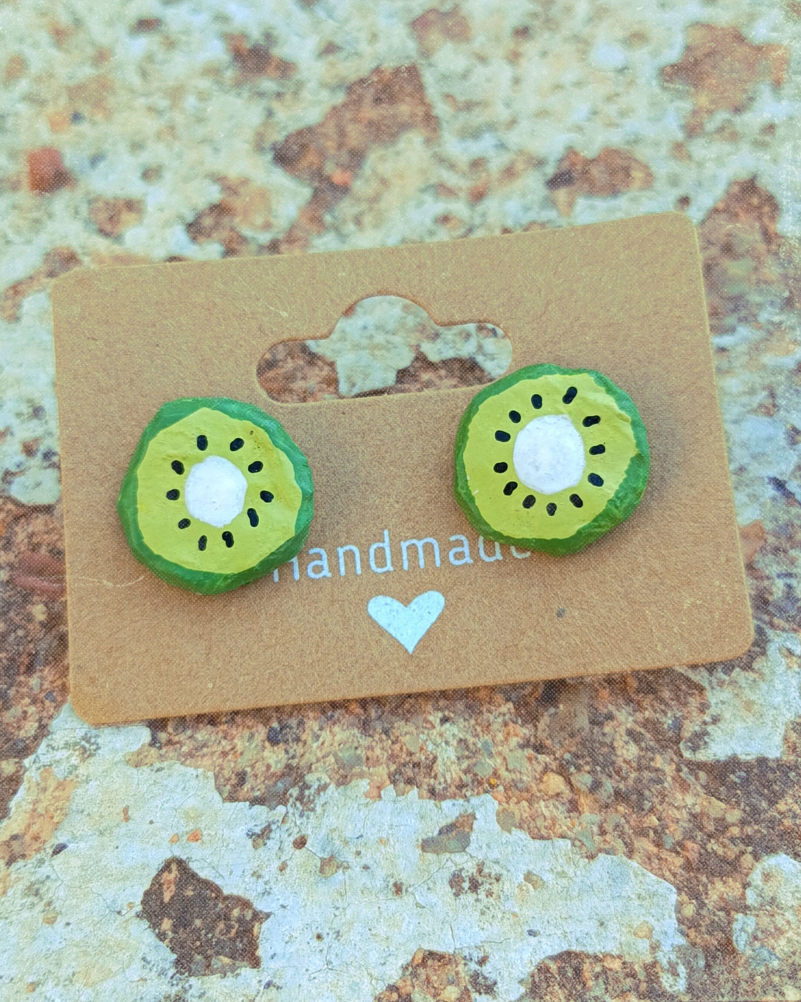 Pendientes infantiles con forma de kiwi sobre packaging de cartón colocados sobre fondo de piedra